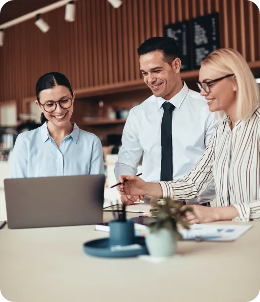 Three colleagues smiling and collaborating around a laptop in a modern office setting.